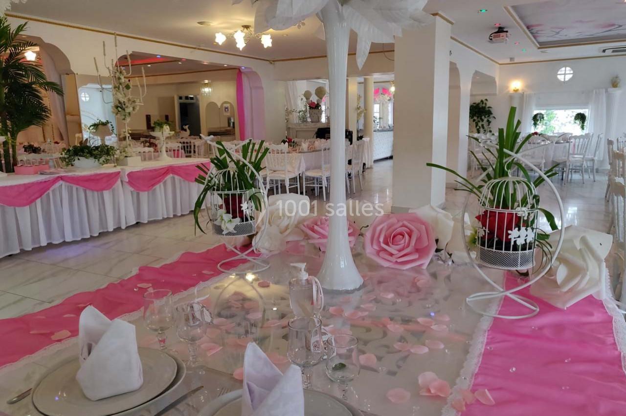Salle de réception décorée avec des nappes roses, des roses artificielles et des tables dressées pour un événement.