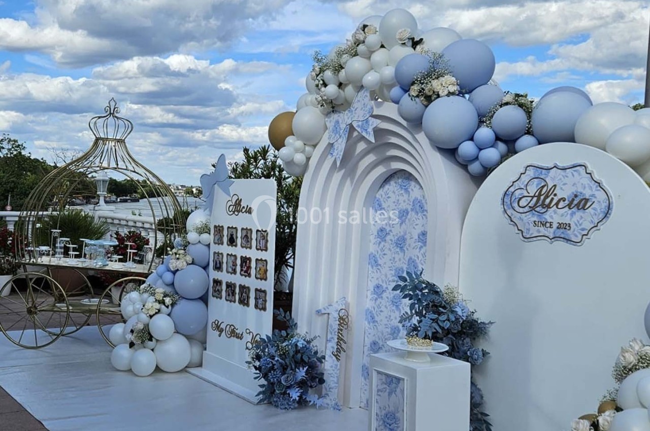 Décoration festive avec arche florale, ballons blancs et bleus, et présentoirs pour une célébration en extérieur.