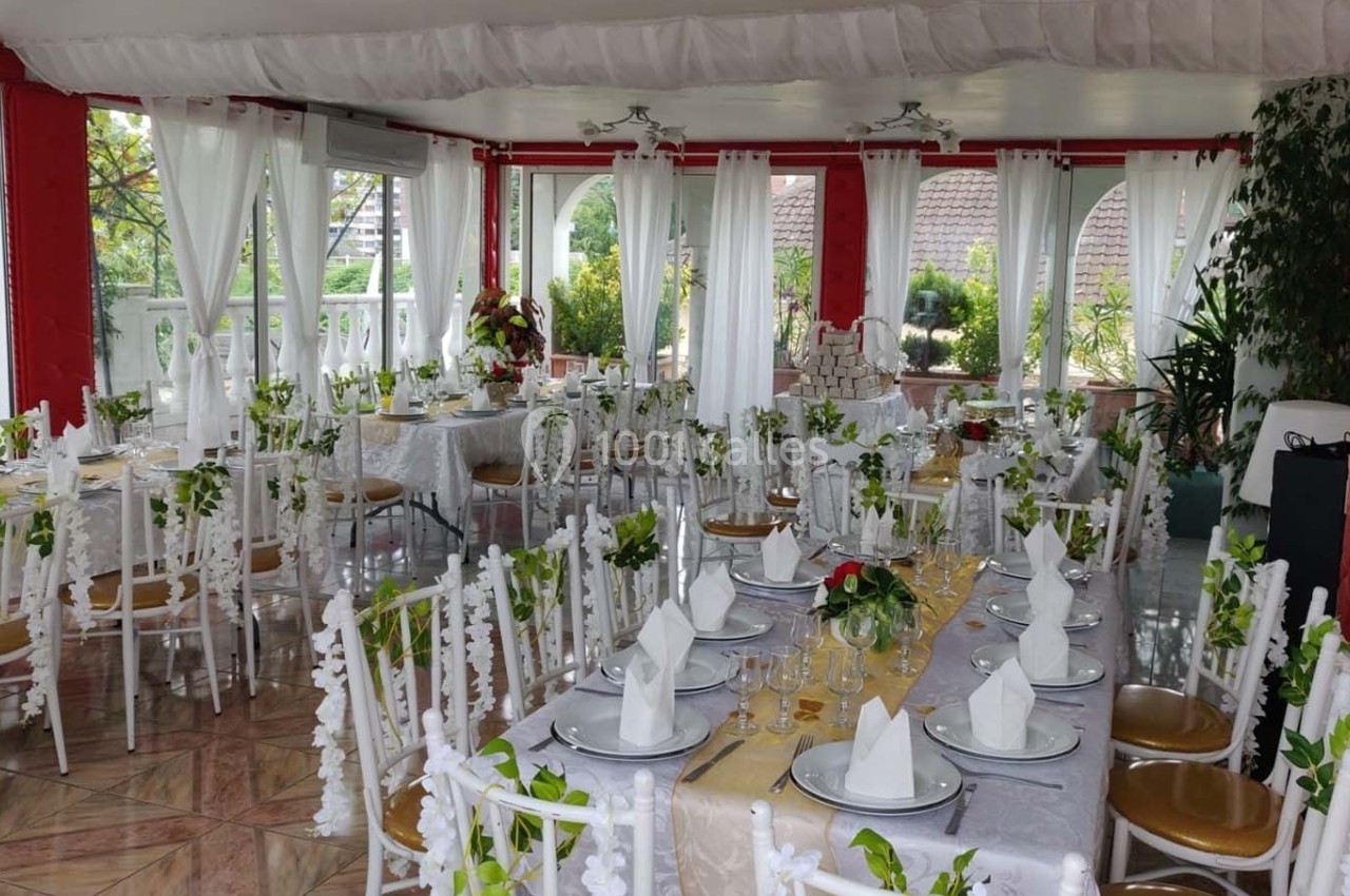 Salle de réception lumineuse décorée pour un événement, avec tables dressées, chaises blanches et ornements floraux.