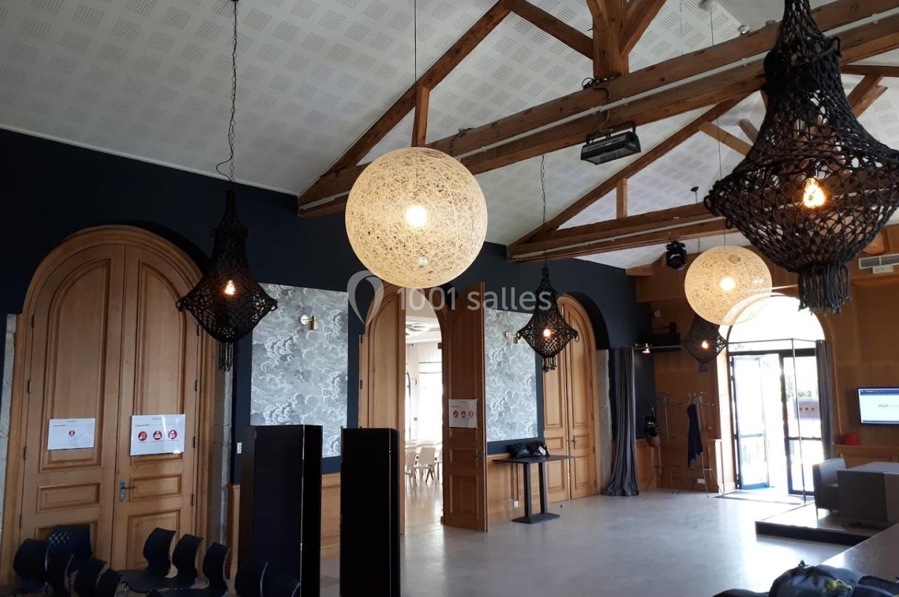Salle avec poutres apparentes, grandes portes en bois, suspensions lumineuses design et chaises disposées en cercle.