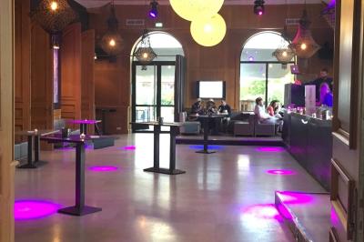 Salle avec un bar, tables hautes et éclairage violet, grandes fenêtres ouvertes sur une terrasse.