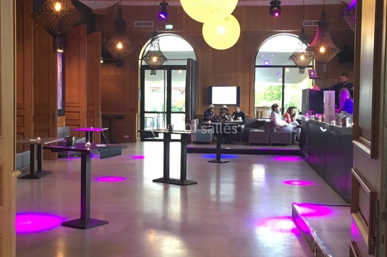 Salle avec un bar, tables hautes et éclairage violet, grandes fenêtres ouvertes sur une terrasse.