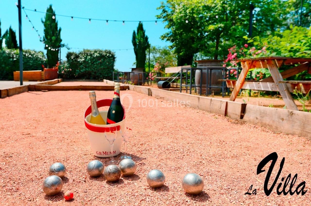 Terrain de pétanque avec des boules en métal, un seau contenant des bouteilles et des tables en bois à l'arrière-plan.