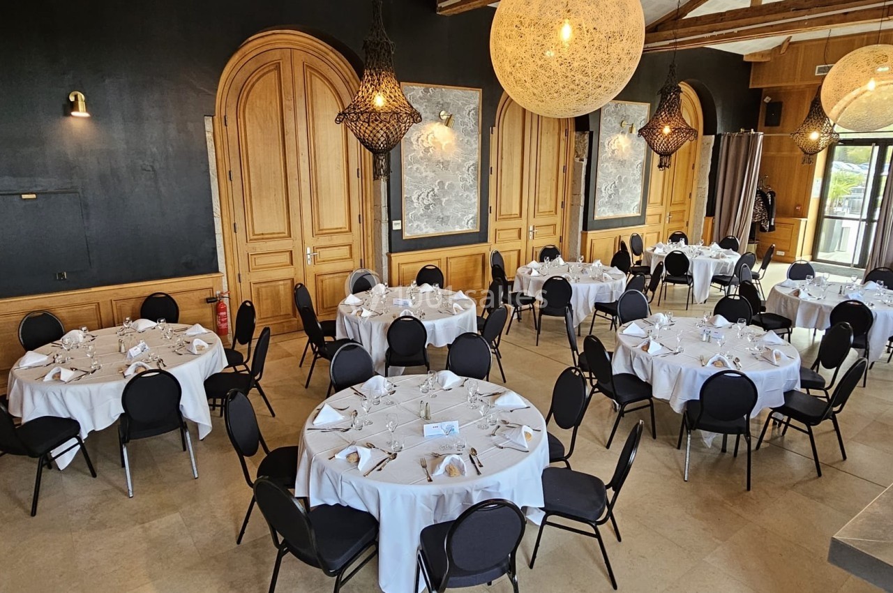 Salle de réception élégante avec tables rondes dressées de nappes blanches, chaises noires et éclairage suspendu.
