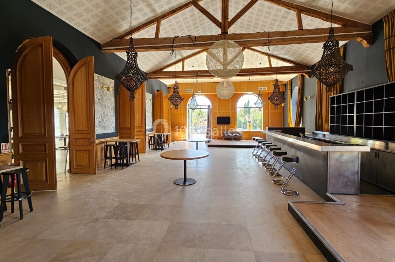 Salle lumineuse avec poutres apparentes, comptoir, tables, chaises hautes et grandes fenêtres donnant sur l'extérieur.