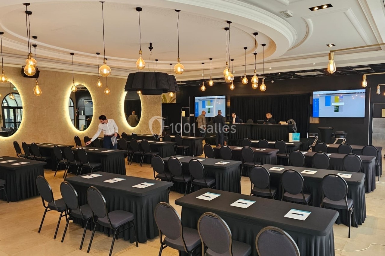 Salle de conférence avec des chaises alignées, des tables noires, des écrans en arrière-plan et des lumières suspendues.