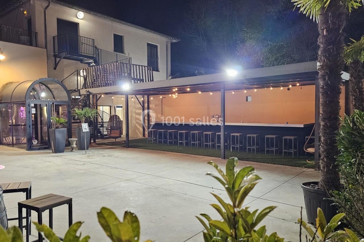 Cour extérieure éclairée la nuit avec un bar couvert, des plantes et des bâtiments en arrière-plan.