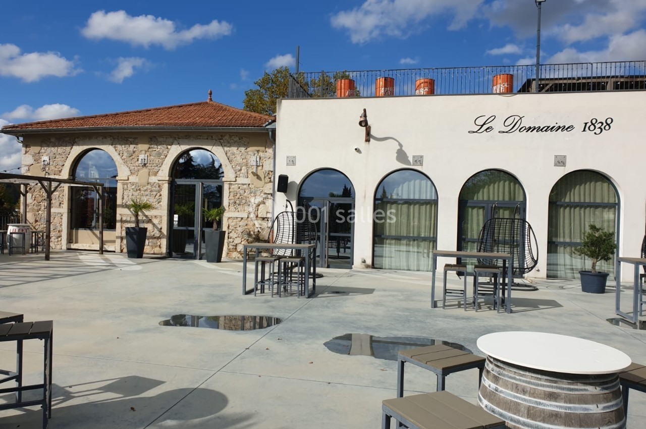 Terrasse extérieure d'un bâtiment en pierre avec des arches vitrées, tables et chaises sous un ciel partiellement nuageux.