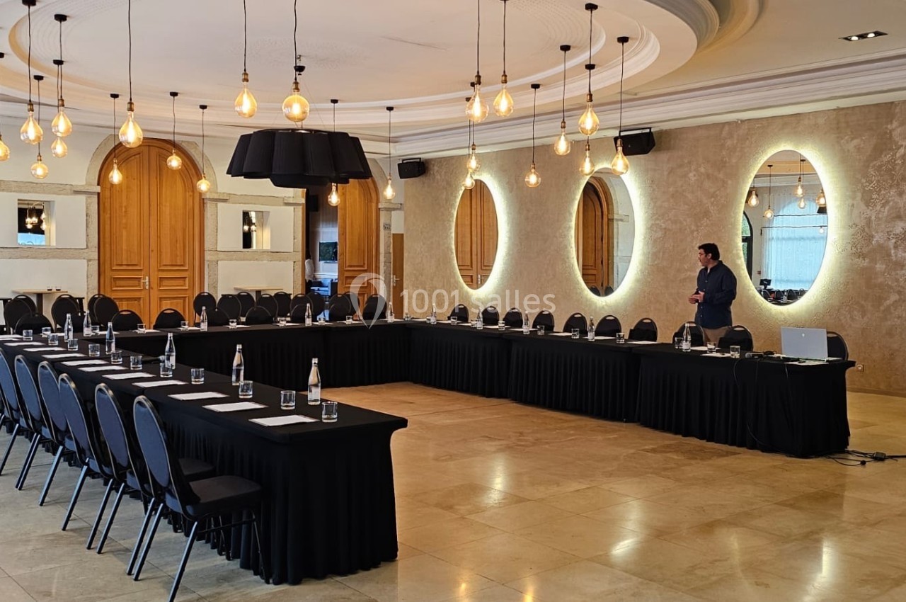 Salle de réunion élégante avec tables disposées en U, chaises alignées, éclairage suspendu et un homme debout à droite.