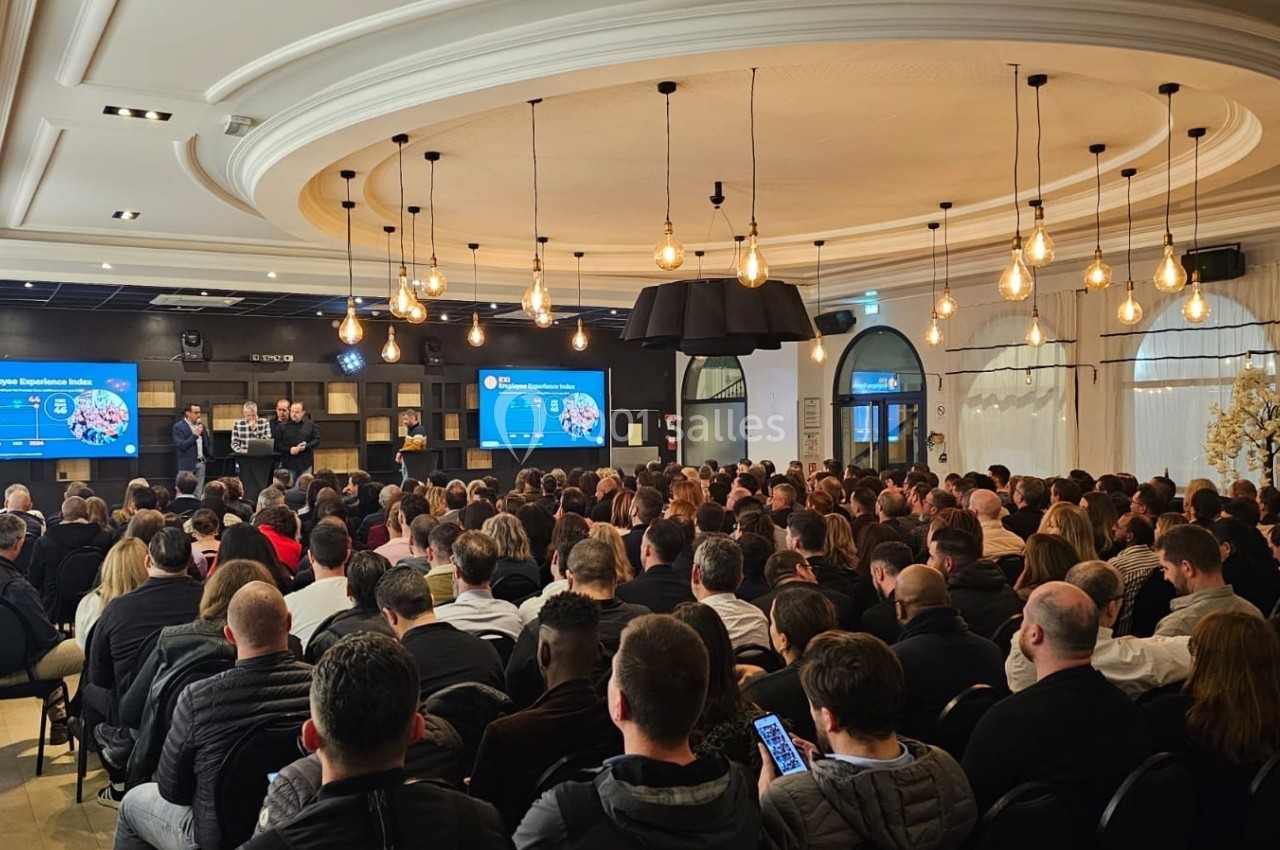 Salle comble avec un public attentif lors d'une présentation sur scène, écrans affichant des diapositives en arrière-plan.