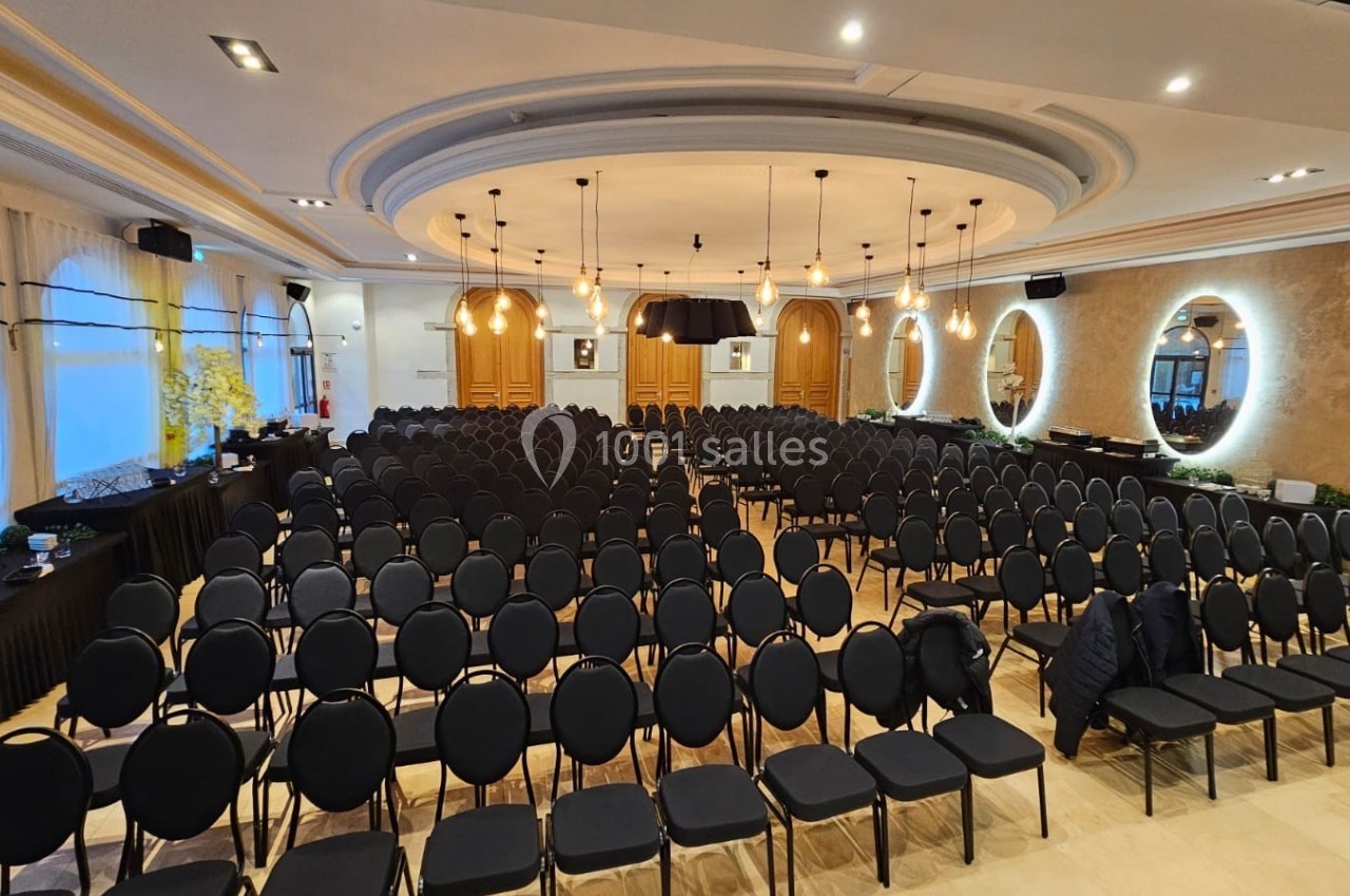 Salle de conférence avec des rangées de chaises noires alignées, éclairée par des lustres et des appliques murales.