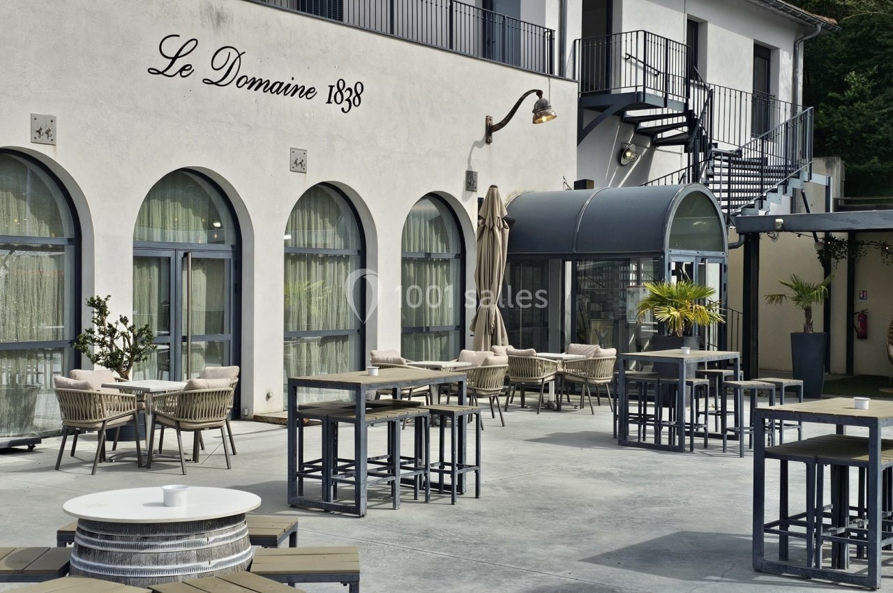 Terrasse extérieure avec tables, chaises et parasols devant un bâtiment aux grandes fenêtres et escaliers métalliques.