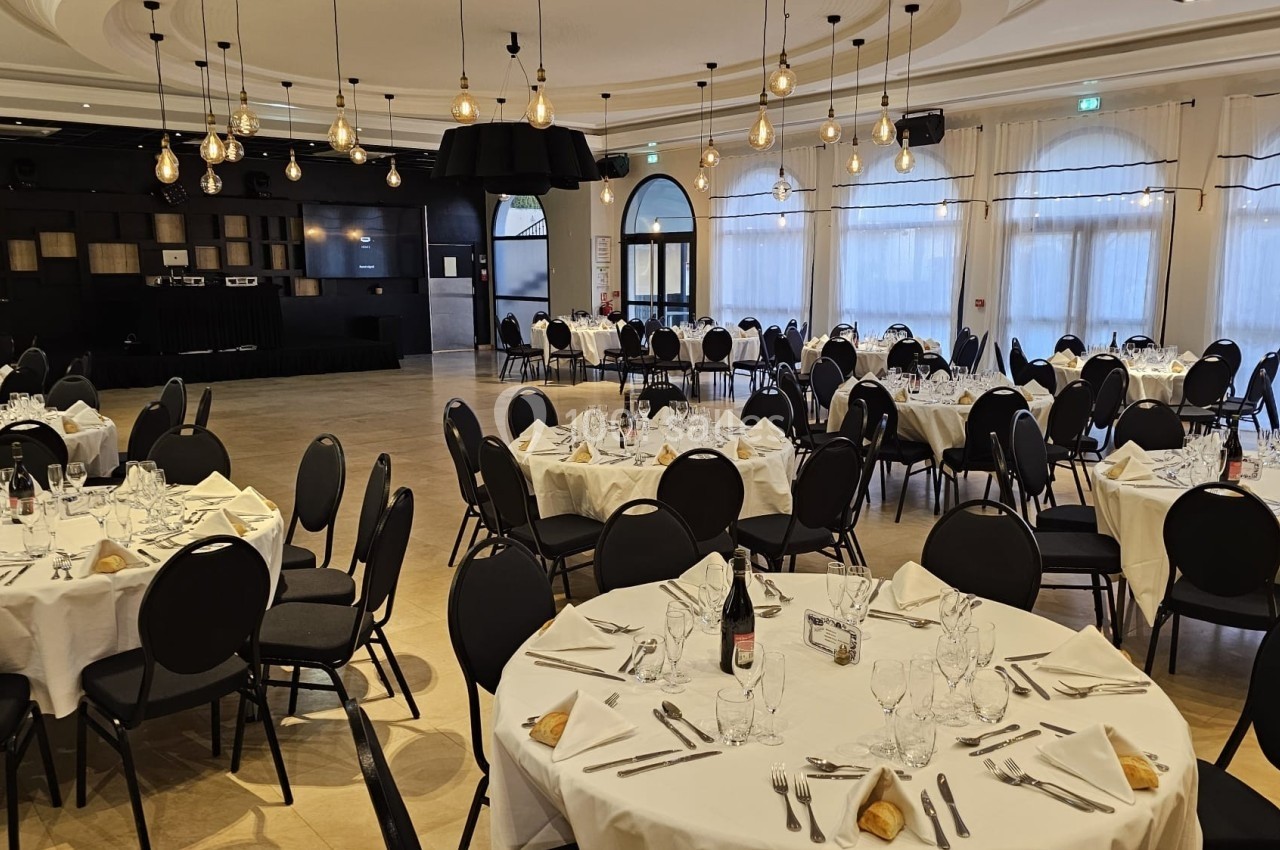 Salle de réception avec tables rondes dressées, nappes blanches, chaises noires et éclairage suspendu.