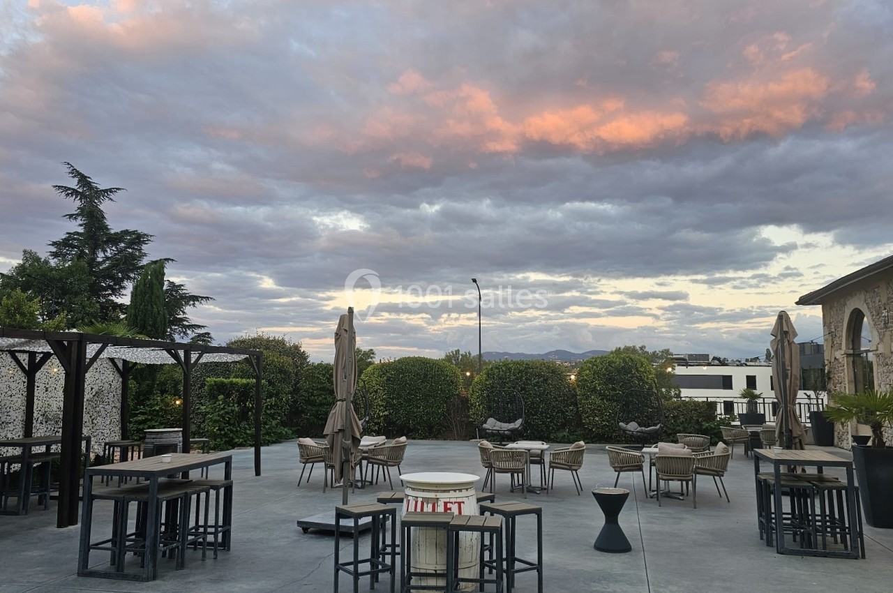 Terrasse extérieure avec tables, chaises et parasols, entourée de verdure, sous un ciel nuageux au coucher du soleil.