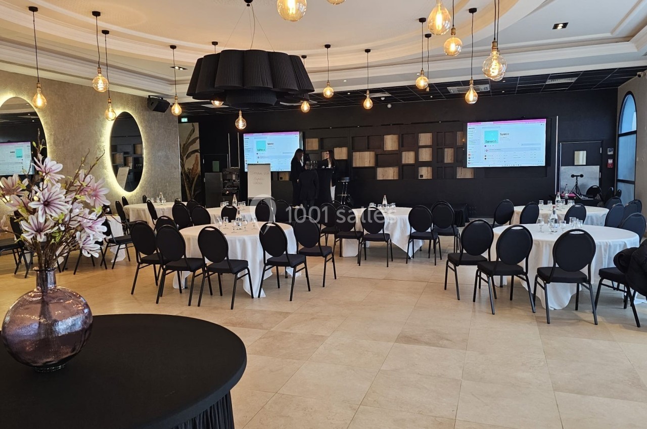 Salle de conférence lumineuse avec tables rondes, chaises noires, écrans de présentation et décoration florale.