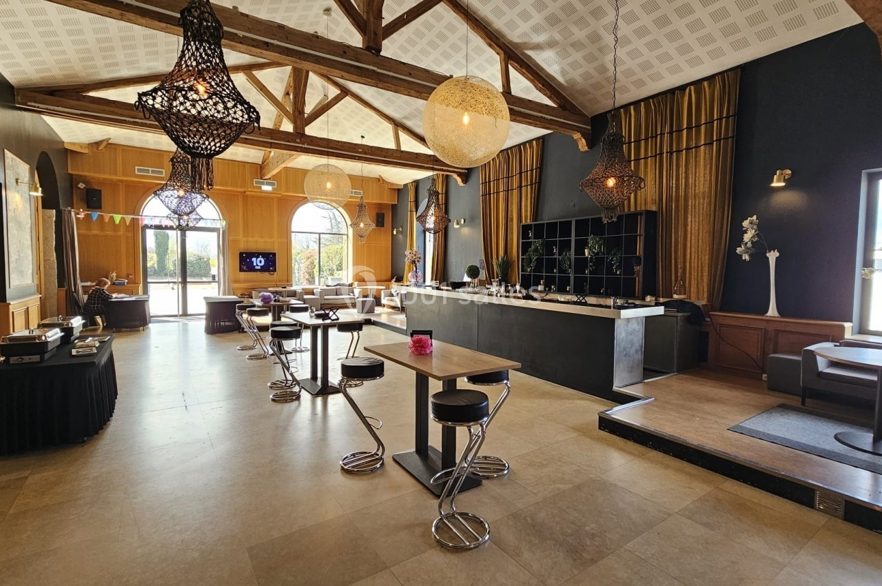 Salle lumineuse avec tables hautes, chaises modernes, grandes fenêtres, poutres apparentes et luminaires suspendus.