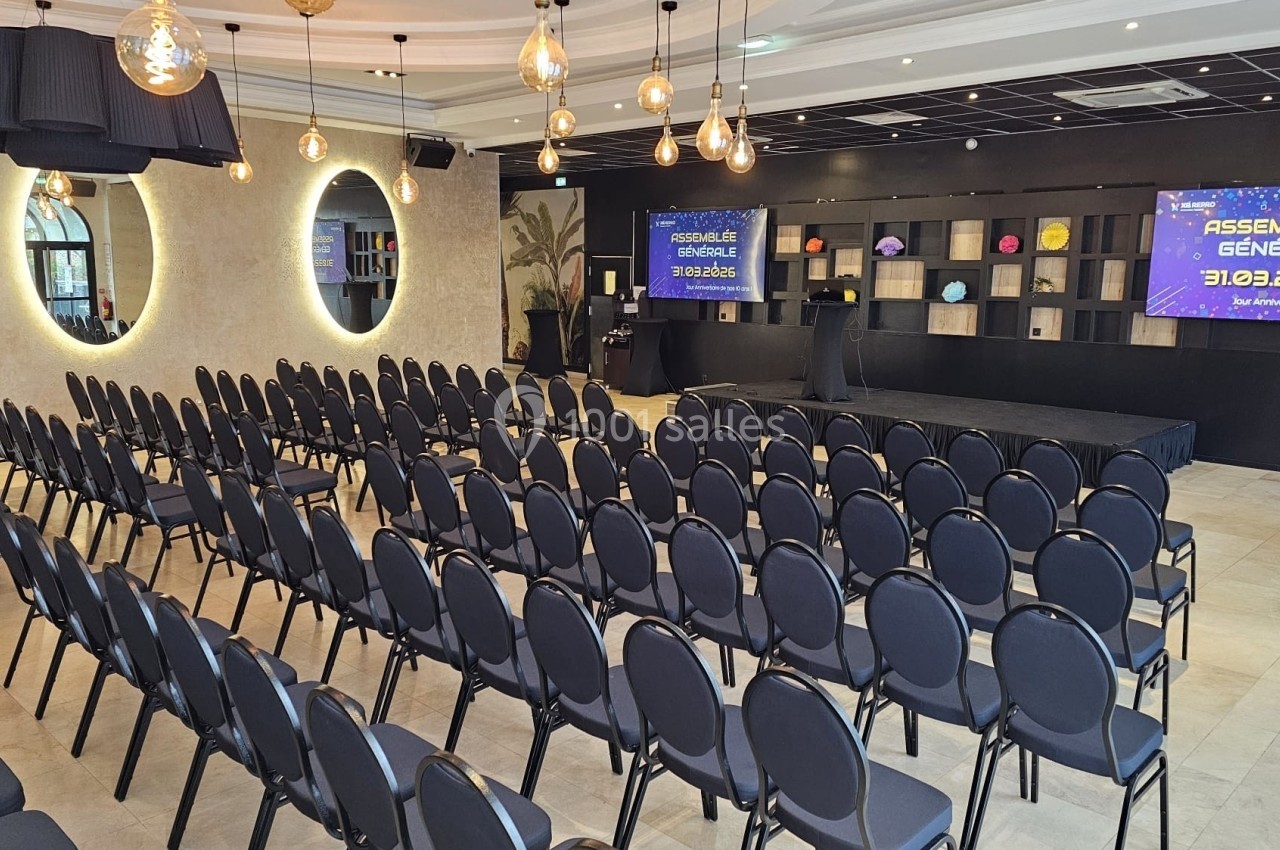 Salle de conférence aménagée avec des rangées de chaises noires, une scène et des écrans affichant ’Assemblée Générale 31.03…