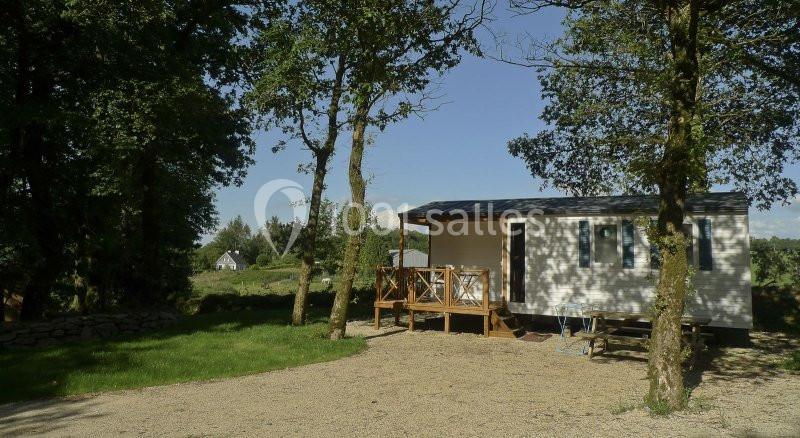 Mobil-home blanc avec terrasse en bois, entouré d'arbres, sur un terrain ensoleillé avec une maison au loin.