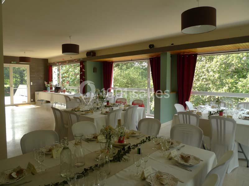 Salle lumineuse décorée pour un repas, avec tables dressées, nappes blanches, chaises et grandes baies vitrées donnant sur…
