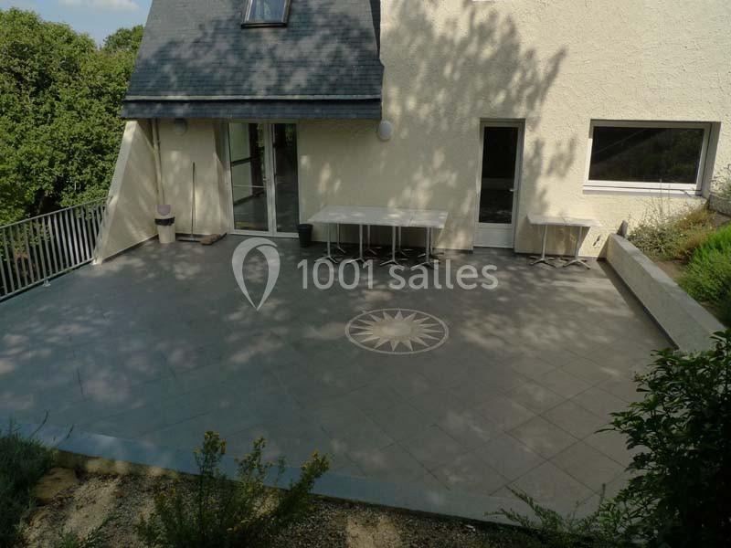 Grande terrasse en carrelage gris avec une rosace centrale, attenante à une maison claire et entourée de verdure.