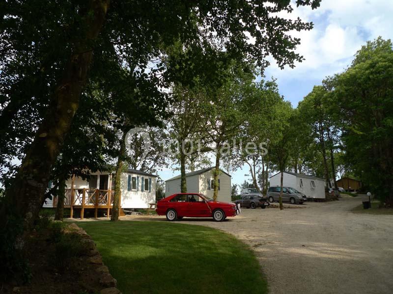 Alignement de mobil-homes dans un camping arboré, avec une voiture rouge garée sur une pelouse.