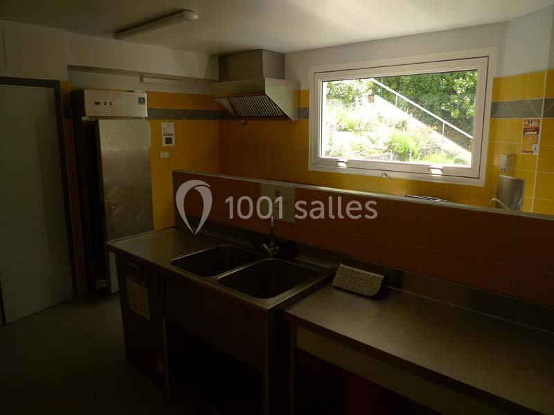 Cuisine équipée avec évier double, plan de travail en inox, fenêtre laissant entrer la lumière naturelle et réfrigérateur.