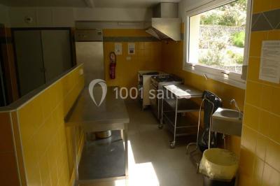 Cuisine professionnelle avec murs jaunes, plan de travail en inox, évier, cuisinière, fenêtre et équipements de rangement.