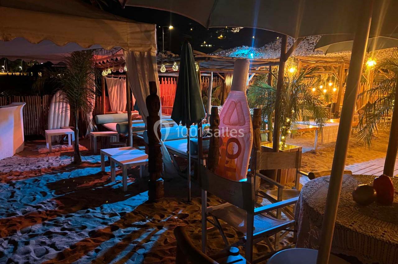 Terrasse de plage éclairée la nuit, avec chaises, tables, parasols et décorations en bois sous des lumières tamisées.