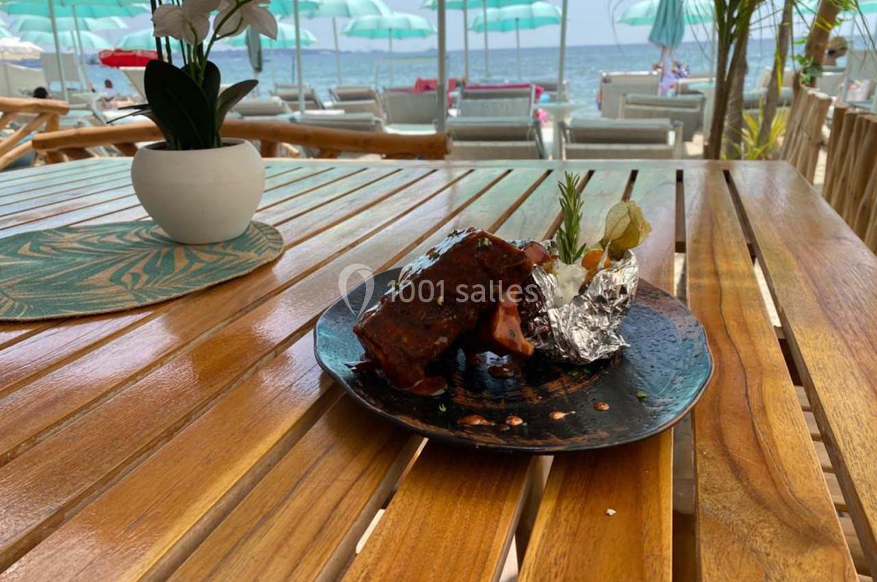Assiette de côtes grillées avec garniture, servie sur une table en bois face à une plage avec parasols et transats.