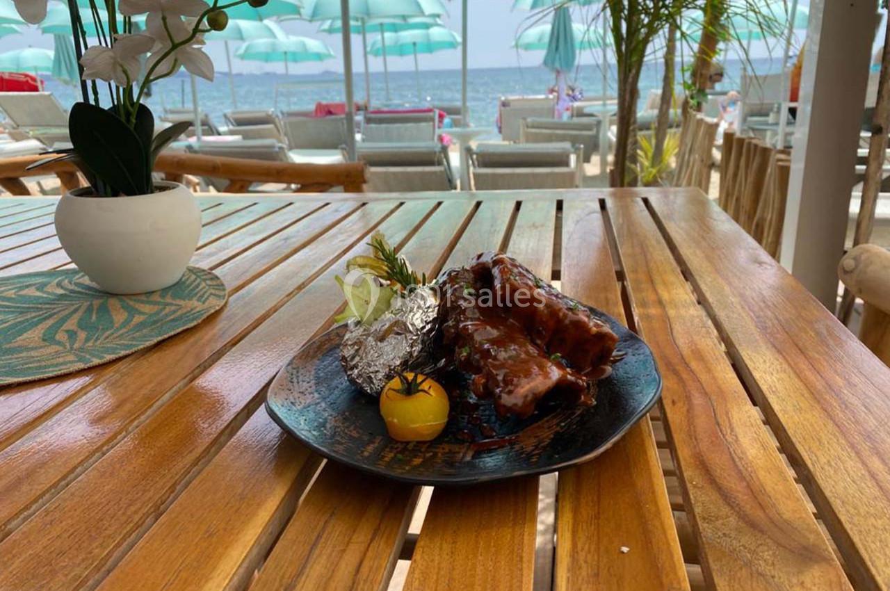 Assiette de côtes levées grillées avec garniture, servie sur une table en bois face à la plage et des parasols.