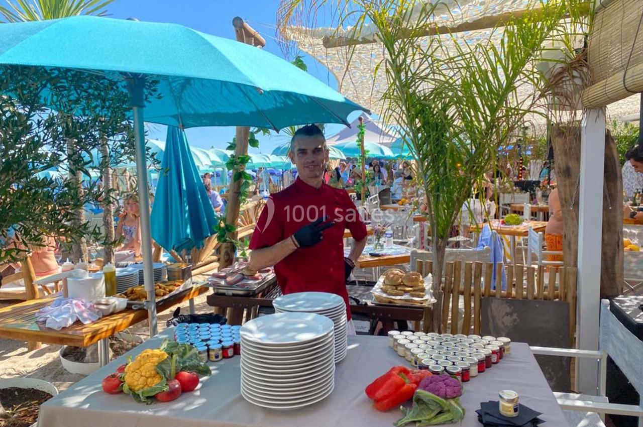 Un serveur devant une table dressée avec des assiettes, légumes et condiments, dans un restaurant en plein air.
