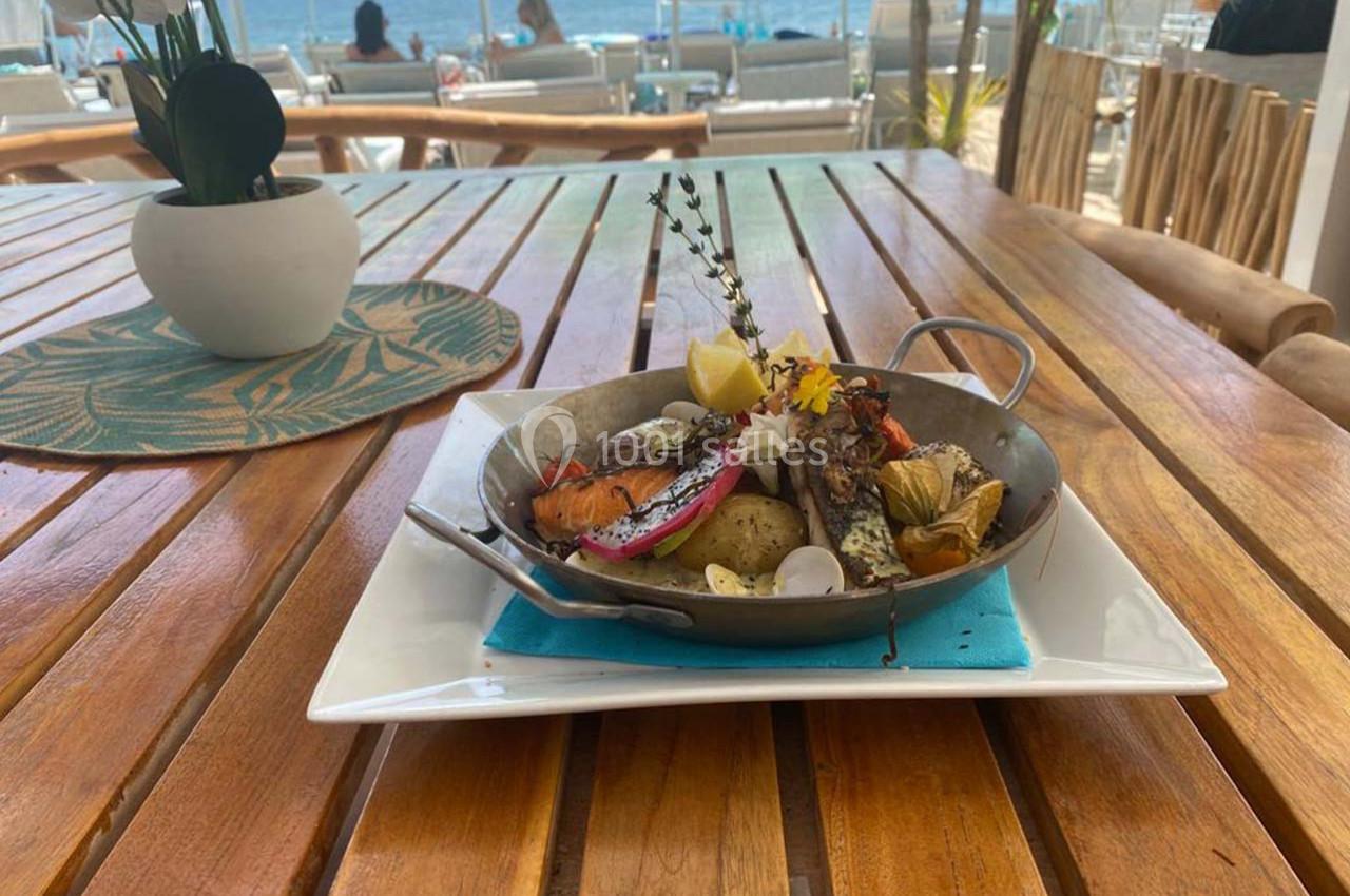 Plat de fruits de mer grillés avec légumes, citron et herbes, servi dans une poêle sur une table en bois près de la plage.