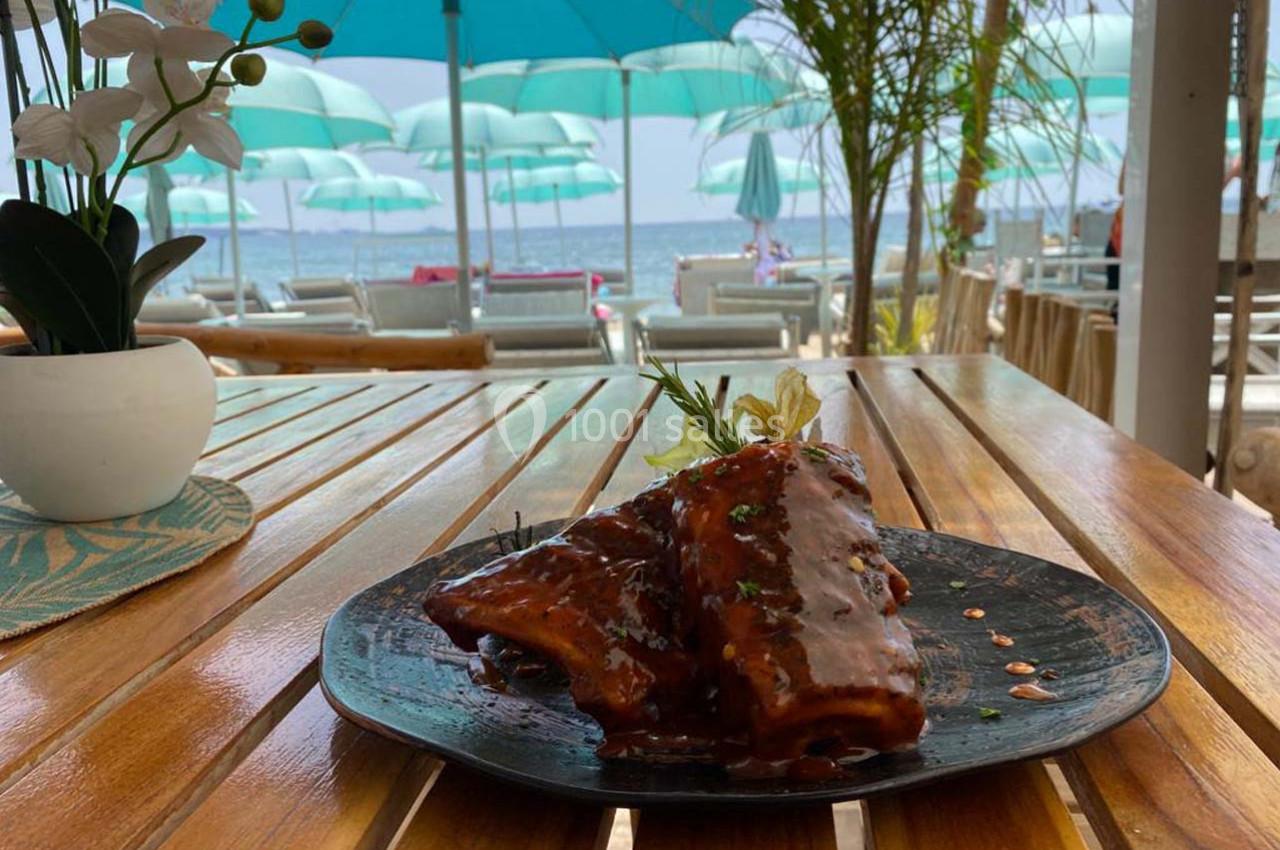 Assiette de travers de porc caramélisés sur une table en bois, avec vue sur une plage et des parasols turquoise.