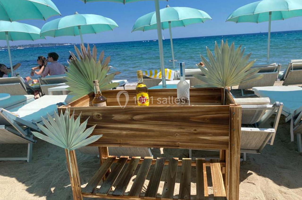 Chariot en bois avec bouteilles, placé sur une plage sous des parasols turquoise près de transats face à la mer.