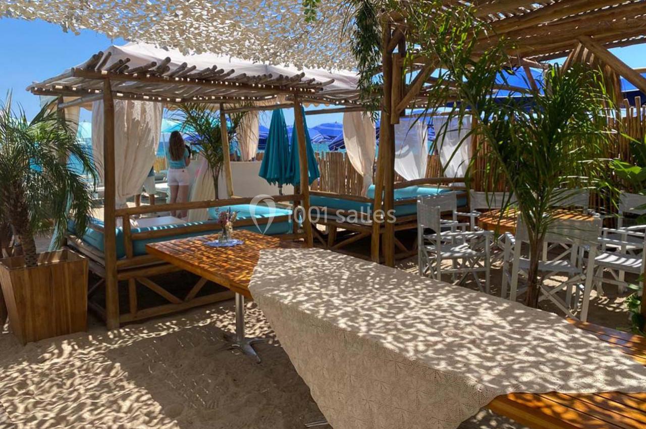 Espace de détente sur une plage avec lits baldaquins, table en bois, chaises et ombrage partiel sous un filet.