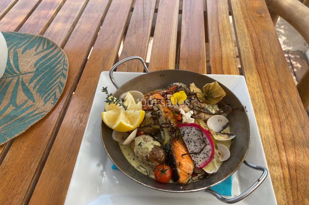Plat de fruits de mer et légumes grillés dans une poêle, garni de citron, herbes et fleurs comestibles sur une table en bois.