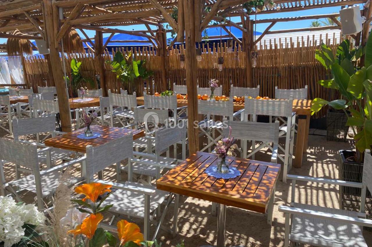Terrasse en bois avec tables et chaises blanches, décorée de plantes et fleurs, sous une pergola ombragée.