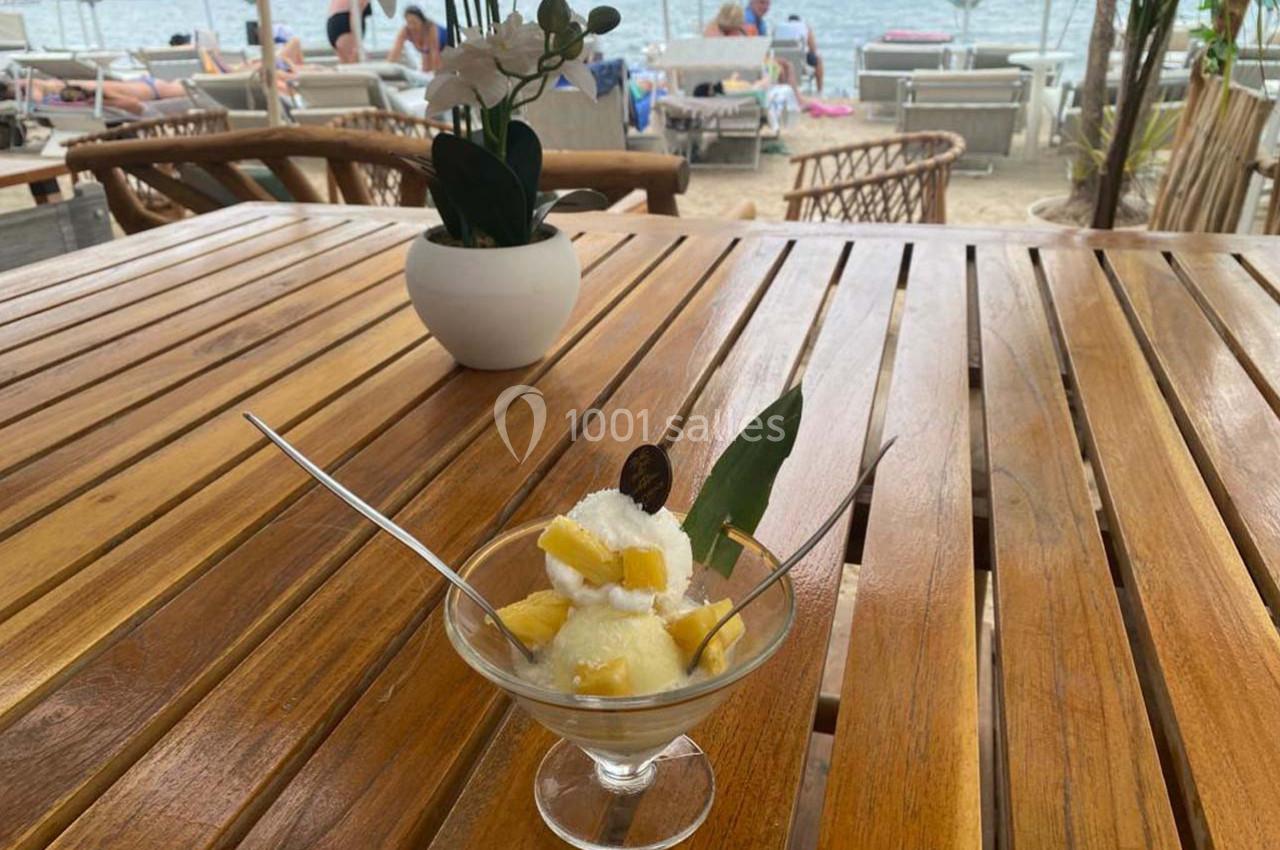 Coupe de glace garnie de morceaux de mangue et d'une feuille, posée sur une table en bois face à une plage.