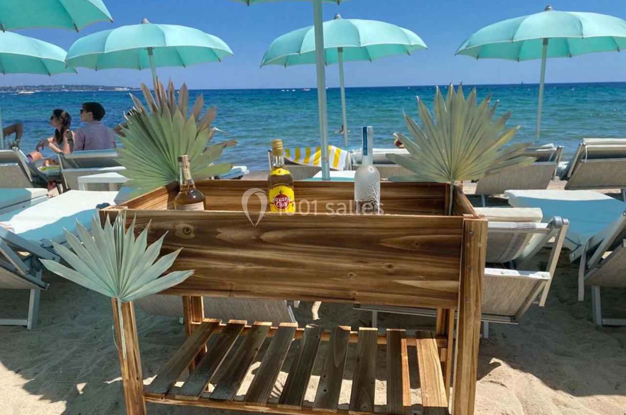 Chariot en bois avec des bouteilles d'alcool, placé sur une plage avec parasols turquoise et vue sur la mer.