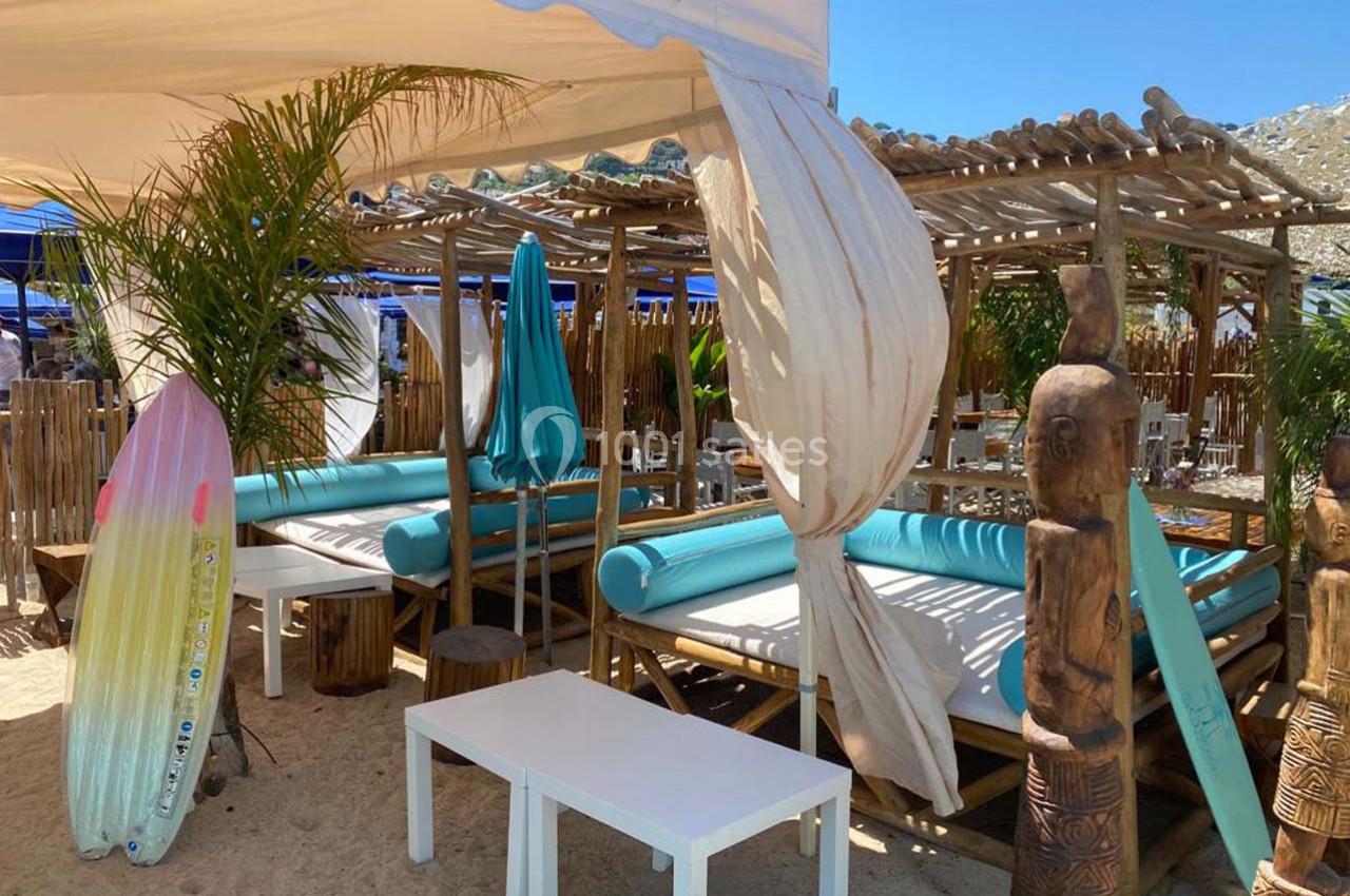 Terrasse en bois avec lits de plage turquoise, parasols et décorations tropicales, sur du sable en plein soleil.