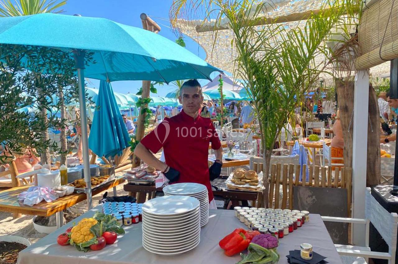 Un cuisinier en tenue rouge devant une table dressée avec des assiettes et des ingrédients, dans un cadre extérieur…