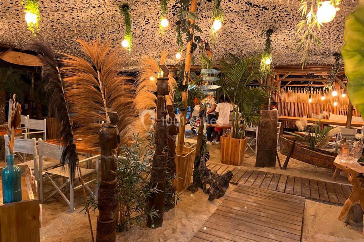 Espace extérieur décoré de plantes, bois et lumières suspendues, avec sol en sable et ambiance chaleureuse.