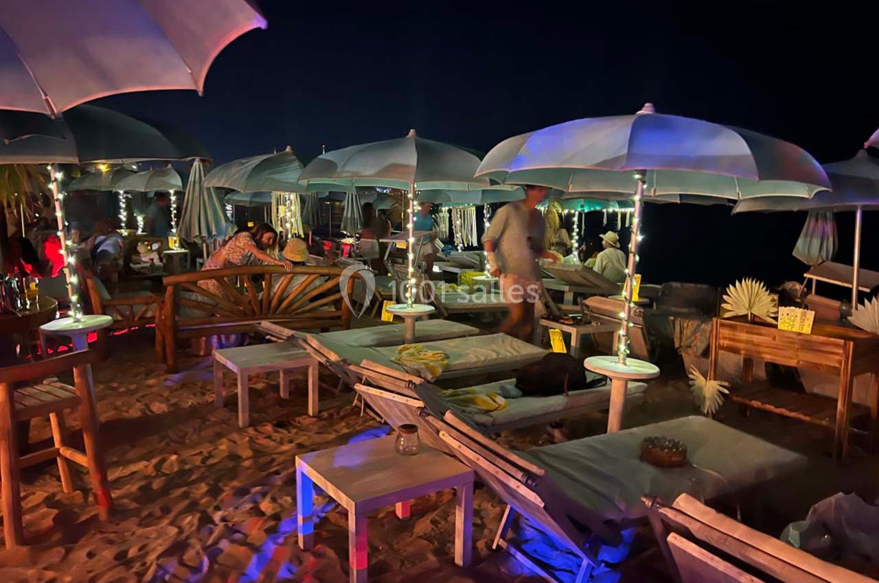 Chaises longues et tables sous des parasols éclairés sur une plage en soirée, ambiance conviviale et détendue.