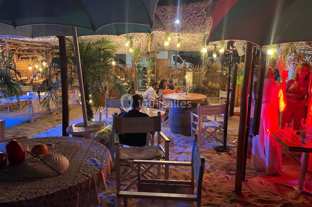 Terrasse de plage éclairée en soirée avec tables, chaises en bois, parasols et ambiance conviviale.