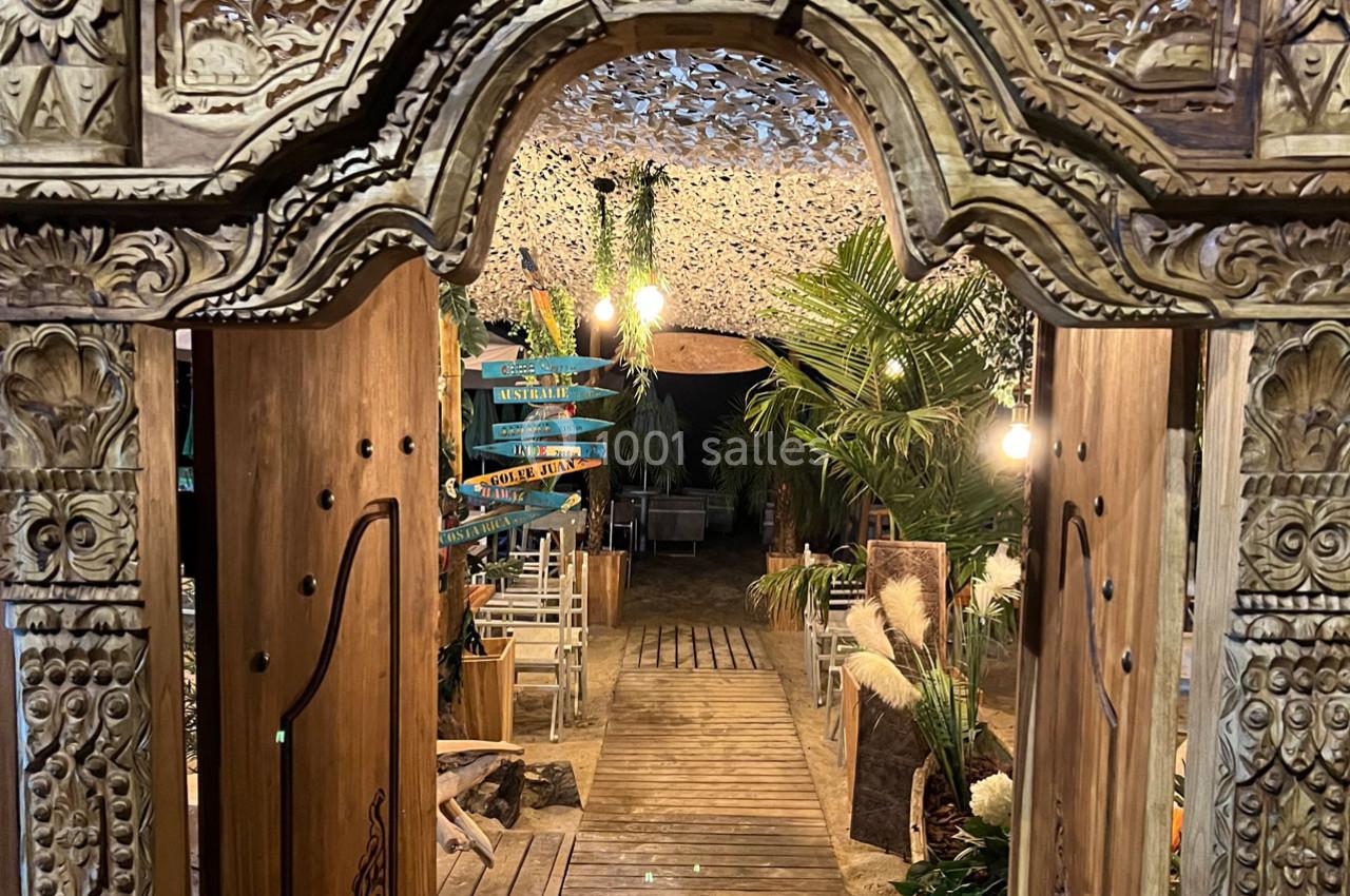 Entrée en bois sculpté menant à un espace extérieur décoré de plantes, lumières suspendues et panneaux directionnels colorés.