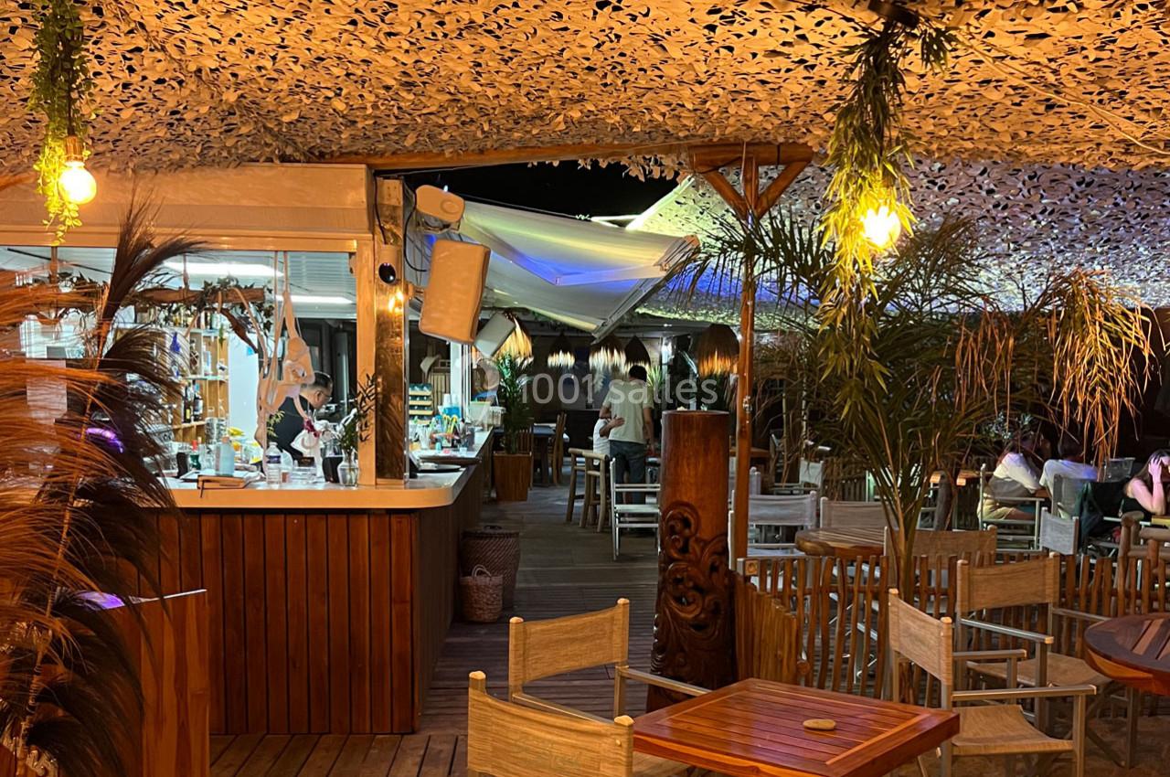 Terrasse extérieure d'un restaurant éclairée en soirée, avec des tables en bois et un bar en arrière-plan.