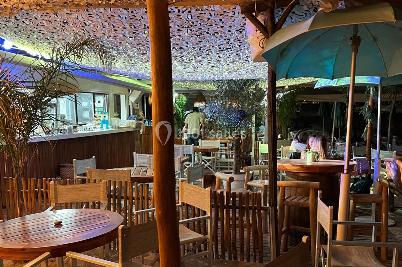 Terrasse d'un restaurant en soirée avec tables en bois, parasols, éclairage tamisé et végétation décorative.