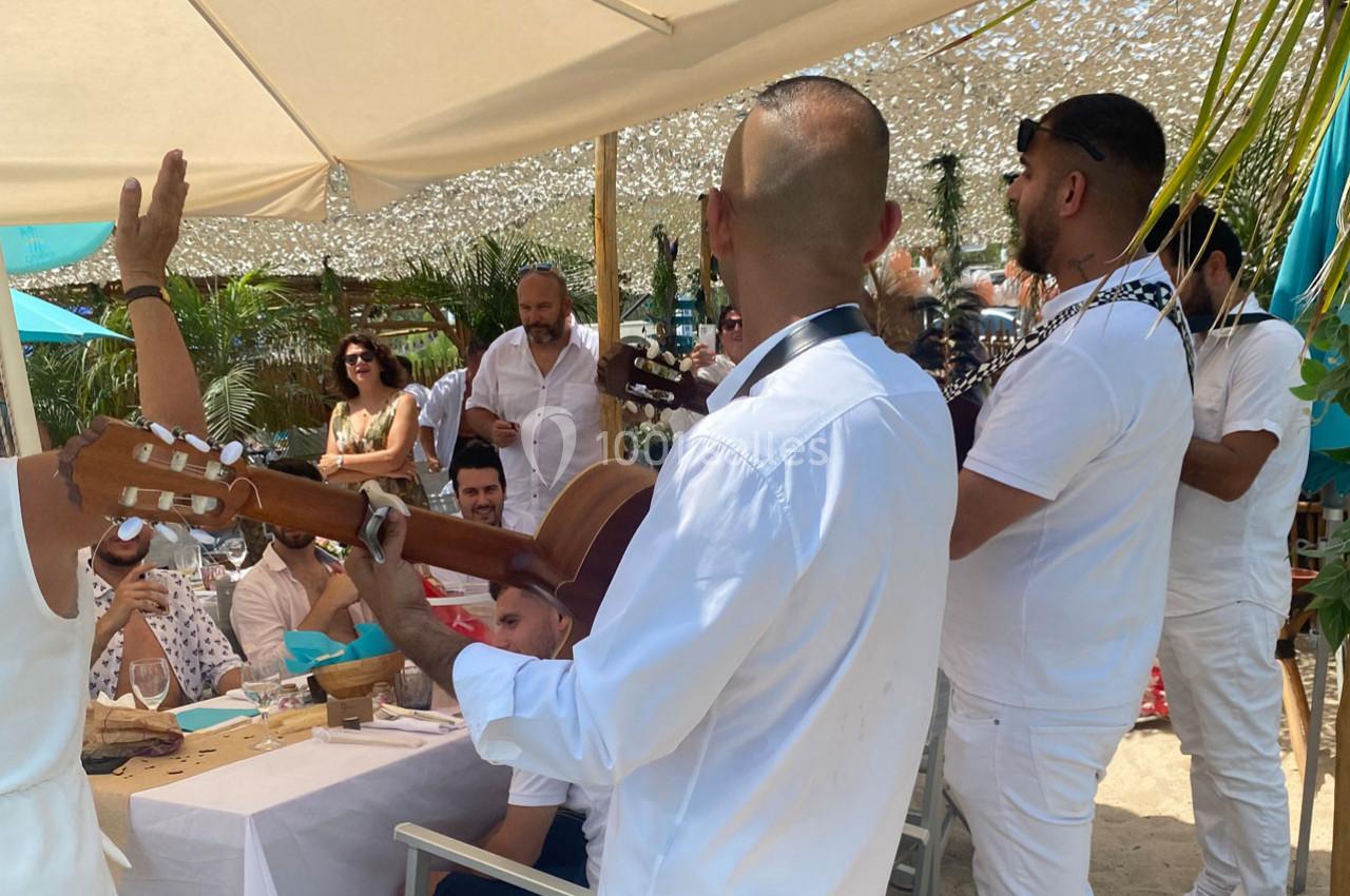 Musiciens jouant de la guitare et animant un repas en plein air sous des parasols, entourés de convives.