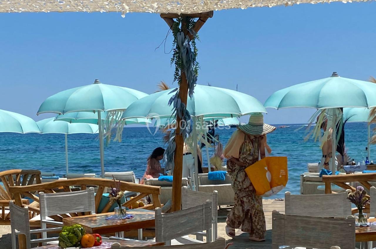 Terrasse d'un restaurant en bord de mer avec parasols turquoise, tables en bois et clients profitant du soleil.