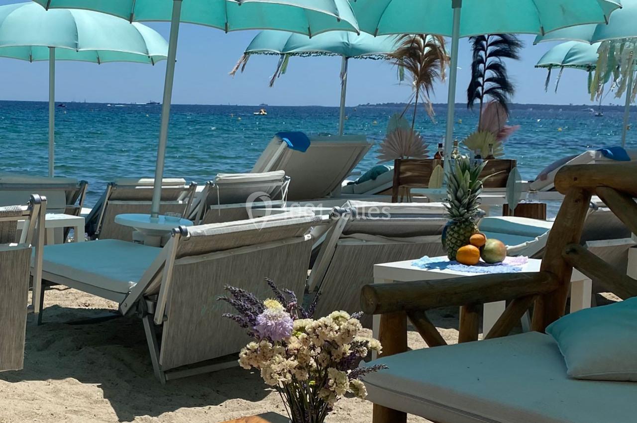 Chaises longues et parasols turquoise disposés sur une plage de sable avec vue sur la mer et des fruits sur une table.